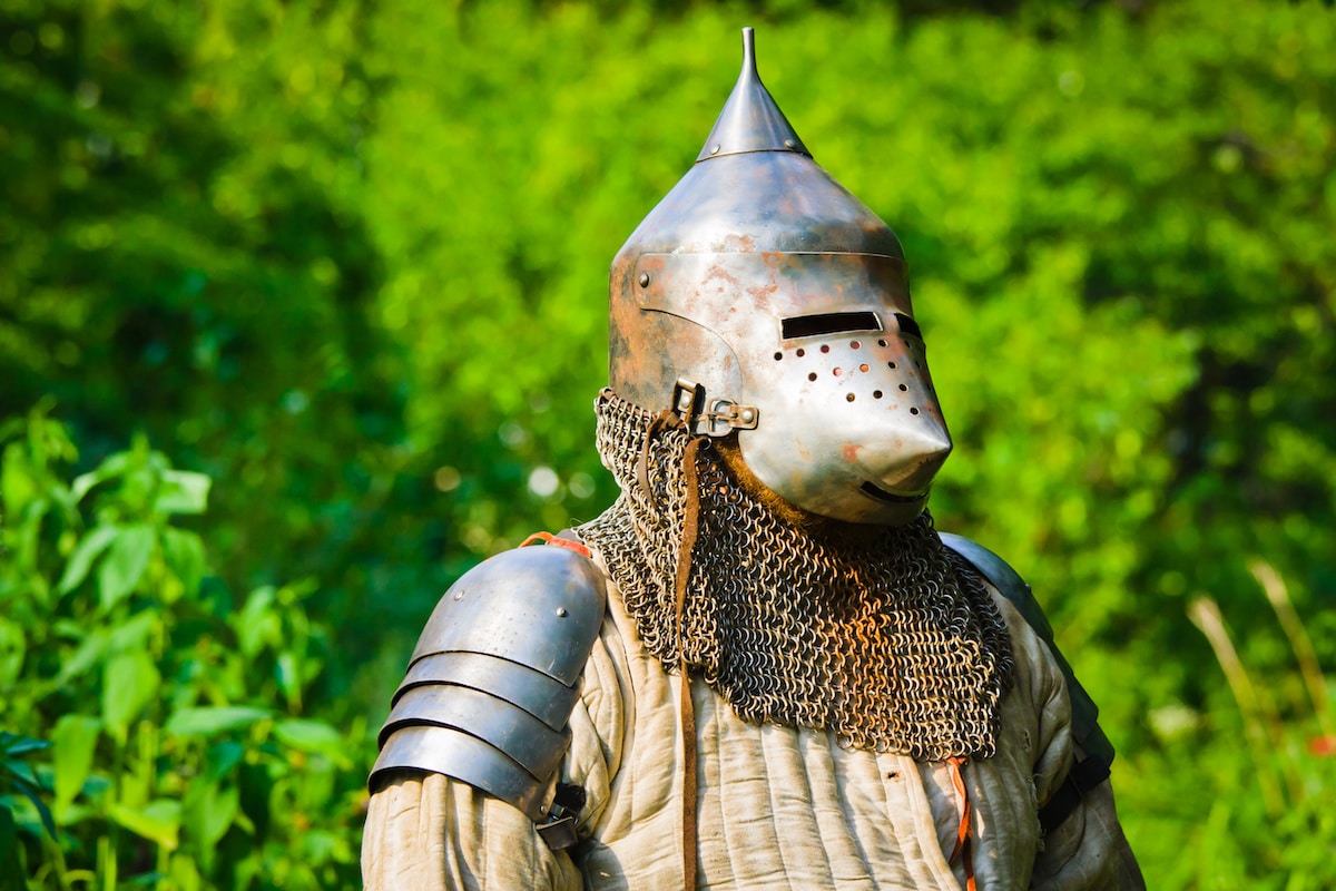 Knight in a rusty helmet and chainmail stands in front of green trees at the Arkansas Renaissance Festival.