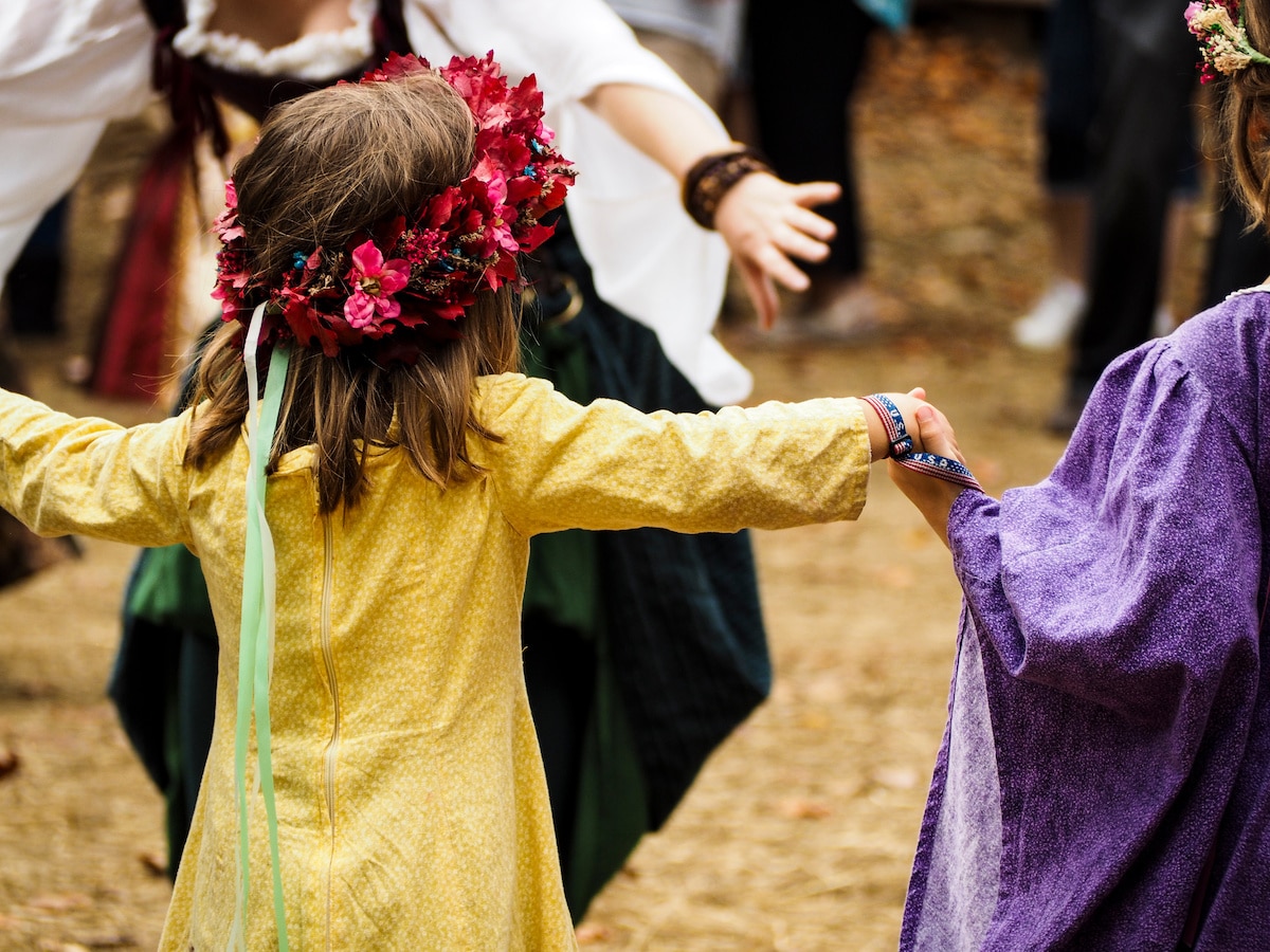 A little girl with a red flower crown is holding hands with the girl next to her and dancing in a circle at the Arkansas Renaissance Festival.