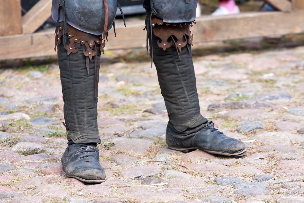 The legs of a man standing on cobblestones wearing leather shoes and medieval knee armor at the Arkansas Renaissance Festival.