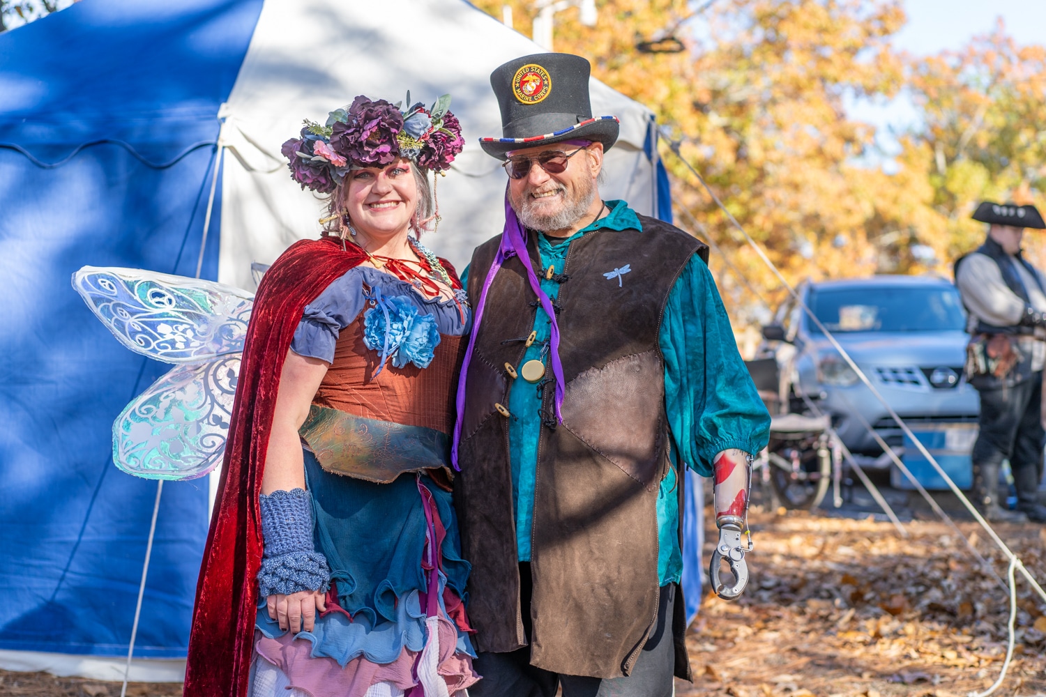 A woman dressed as a flower fairy and a man with a top hat smile for the camera in front of a tent at the Arkansas Renaissance Festival.