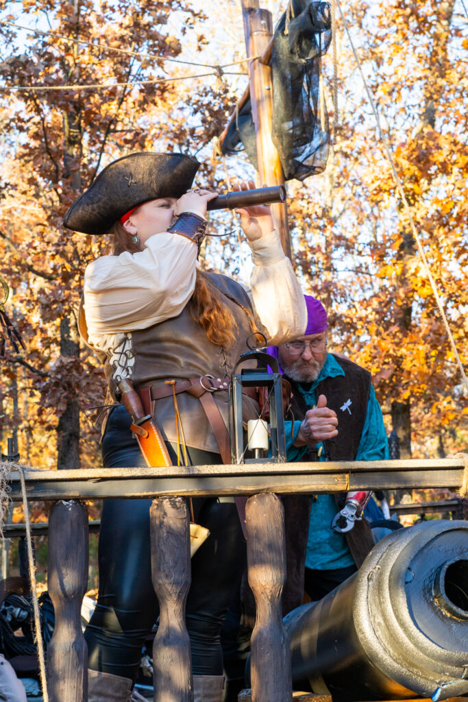 Female pirate cosplayer looks through a spyglass over the side of a wooden pirate ship at the Arkansas Renaissance Festival.